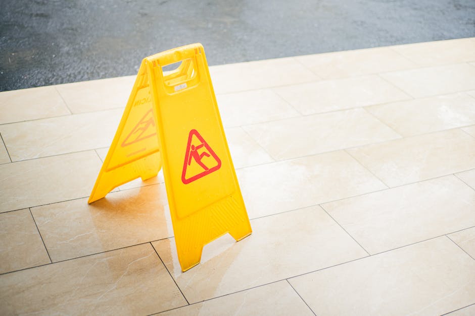 A yellow plastic caution sign with a red warning triangle indicating a wet floor, placed on a beige tiled floor. The sign features an icon of a person slipping, and is positioned in an indoor or outdo