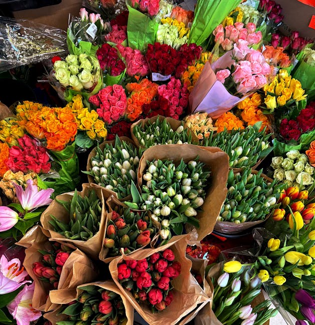A vibrant display of fresh flower bouquets arranged in a florist shop, featuring a variety of blooms including pink lilies, yellow tulips, red roses, white tulips, and green foliage. The bouquets are 
