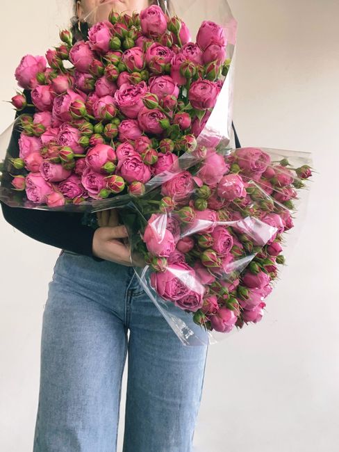 A person holding a large bouquet of fresh pink roses, tightly packed with green buds and lush petals, wrapped in clear cellophane. The bouquet is arranged to showcase the vibrant, fully bloomed flower