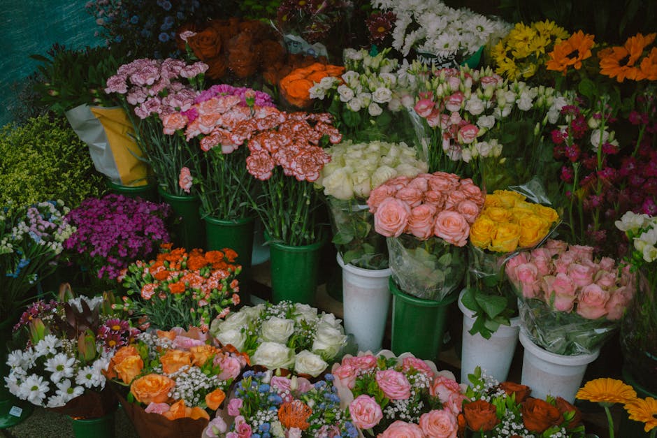 An assortment of fresh floral bouquets and potted flowers displayed on a market stand, featuring a variety of roses, carnations, daisies, and other blossoms in shades of pink, orange, yellow, white, a