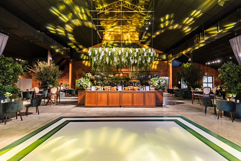 A vibrant indoor event space with a high, dark ceiling illuminated by yellow and white lighting effects. The center features a large wooden buffet table adorned with an abundant floral arrangement of 