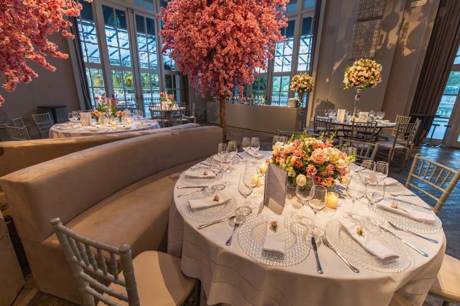 A round table decorated with a large floral centerpiece consisting of pink roses, white carnations, and greenery, arranged in a low, lush bouquet. Surrounding the centerpiece, there are neatly folded 