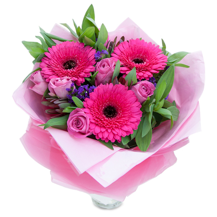 I see this bouquet as a delicate balance of exuberance and softness: three vivid fuchsia Gerbera daisies take centre stage with their broad, radiating petals and deep dark centres, creating an immediate sense of joy. Interspersed among them are roses in a muted lavender-pink-freshly cut, petals curled inward with the tender aroma that only newly opened roses possess-while tiny clusters of purple statice add a papery, airy texture that gives the arrangement a light, garden-like quality. A generous sweep of green foliage, including broad salal-like leaves and slender eucalyptus stems, frames each bloom and lends a cool, leafy contrast to the warm pinks. The bouquet is lovingly wrapped in an inner sheet of soft pale pink tissue paper and overlaid with a translucent, slightly crinkled fuchsia-pink cellophane that glints subtly in soft lighting; the wrapping is folded with intention so the bouquet reads as both polished and intimate. The lower section reveals the top rim of a clear glass vase, suggesting the piece is designed for immediate enjoyment on a mantel or bedside table. Imagining this delivered from a florist serving Primrose Hill and Kentish Town Road to make a morning brighter, the scent feels light and refreshing-roses soft and sweet, gerberas lending a faint grassy note-and the tactile contrast between silky petals and crinkled cellophane invites closer attention. It's a composition that strikes a note of celebration and quiet affection, perfect for anniversaries, birthdays, or a tender 'just because' gift.